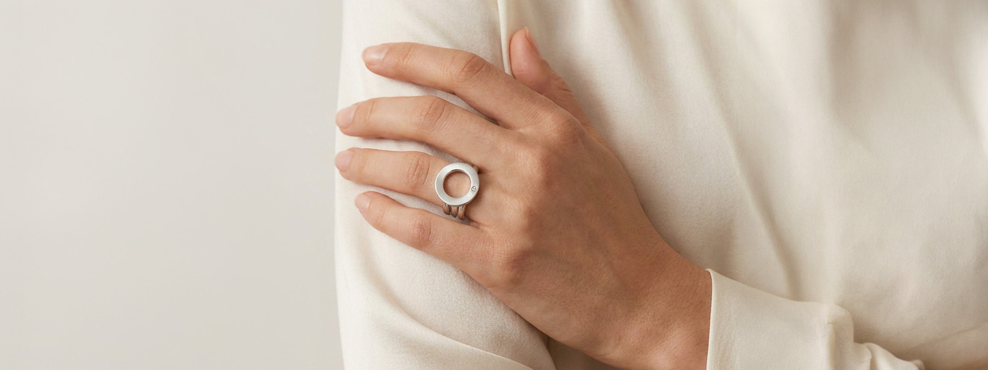 Hand mit silbernem Ring mit rundem Anhänger liegt auf beigem Stoff, weiches Licht im Nahbereich