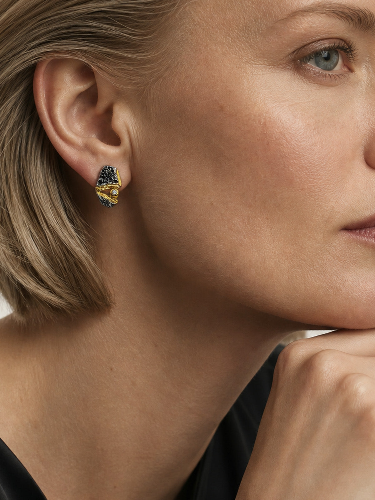 Nahaufnahme eines Ohres mit goldgelbem und schwarzem Schmuck, weichem Gesichtshautdetail und grauem Auge im Hintergrund