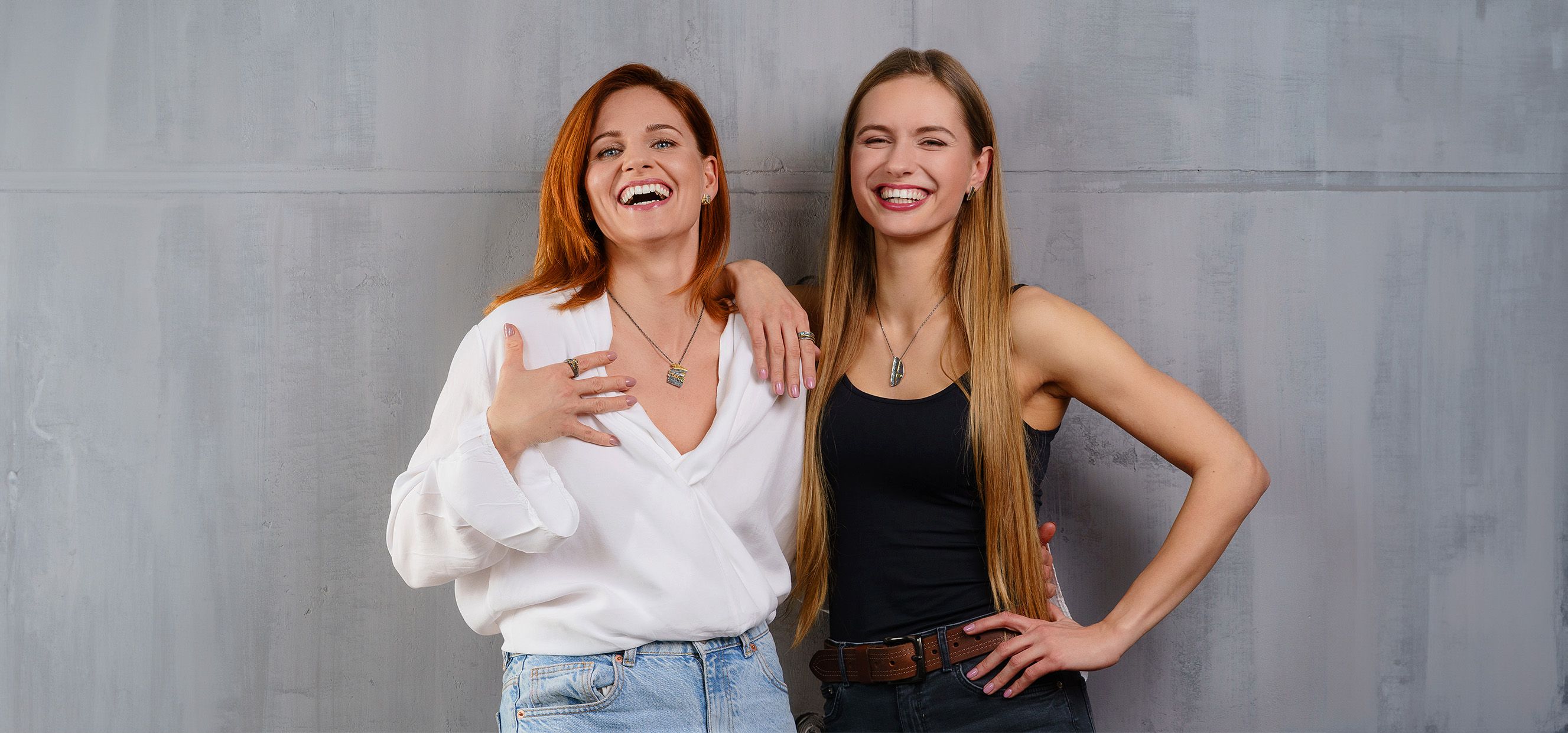 Zwei lächelnde Frauen, die Schmuck tragen, stehen vor einem grauen Hintergrund 